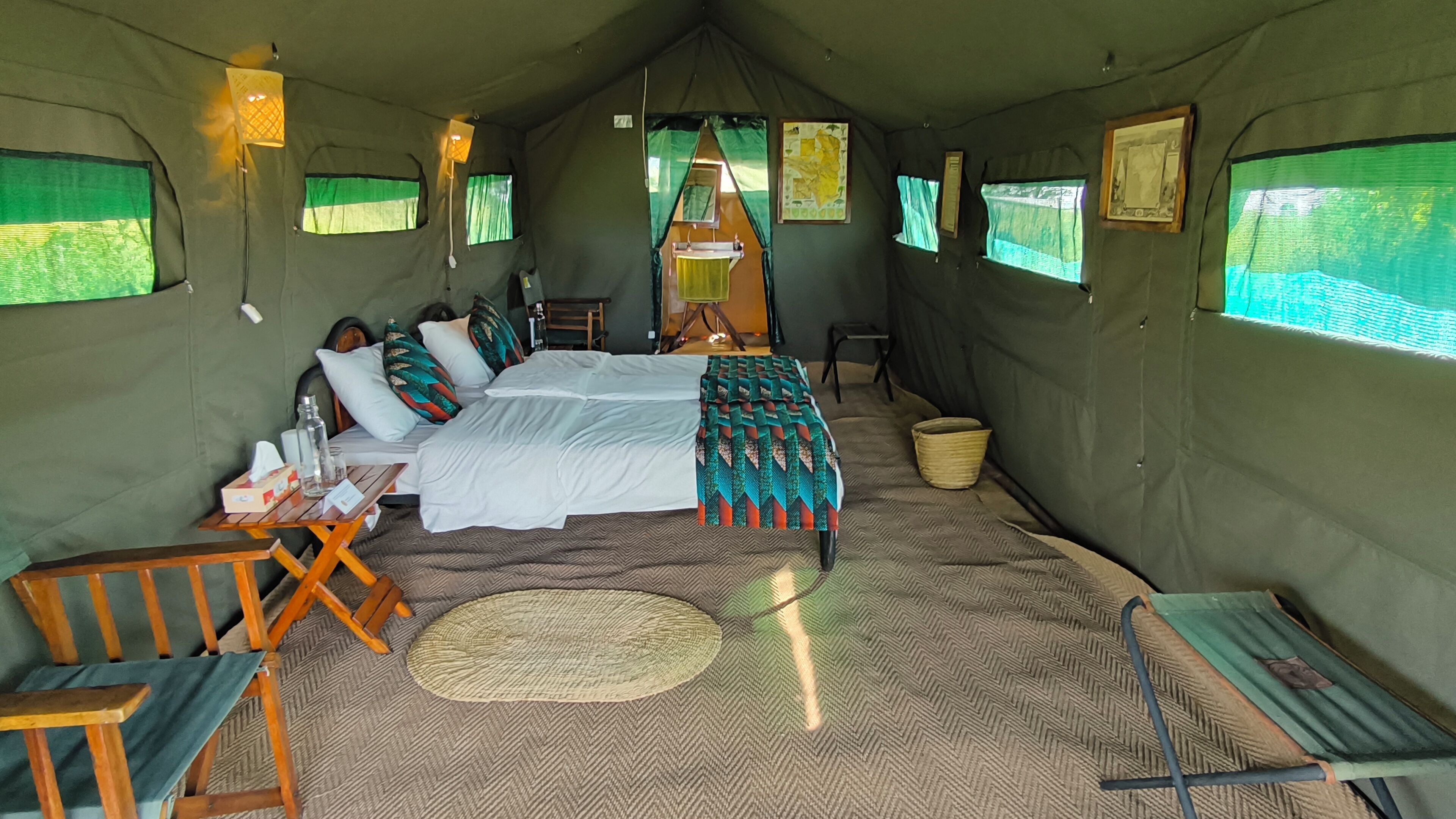 A tent-style room with a bed, a small table, and a chair. There are windows with curtains, a map on the wall, and a woven mat on the floor.