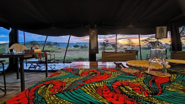 An interior view of a tent with a table set for dining, a large window offering a view of the landscape, and a speaker in the center. - Kananga Special Tented Camp (Serengeti)