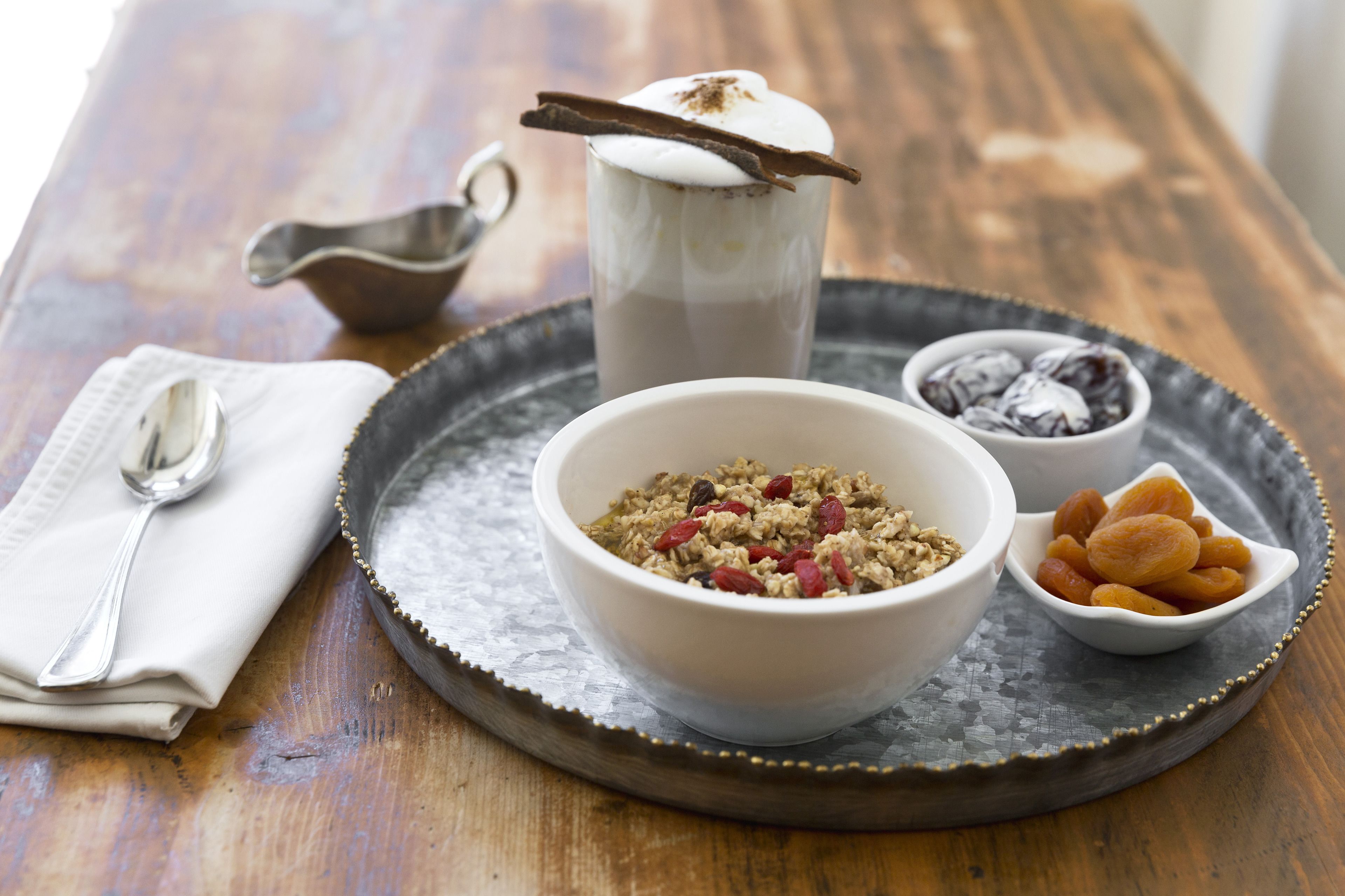 A breakfast setting with a bowl of cereal, a mug of coffee, a small bowl of dried fruits, and a spoon on a tray.
