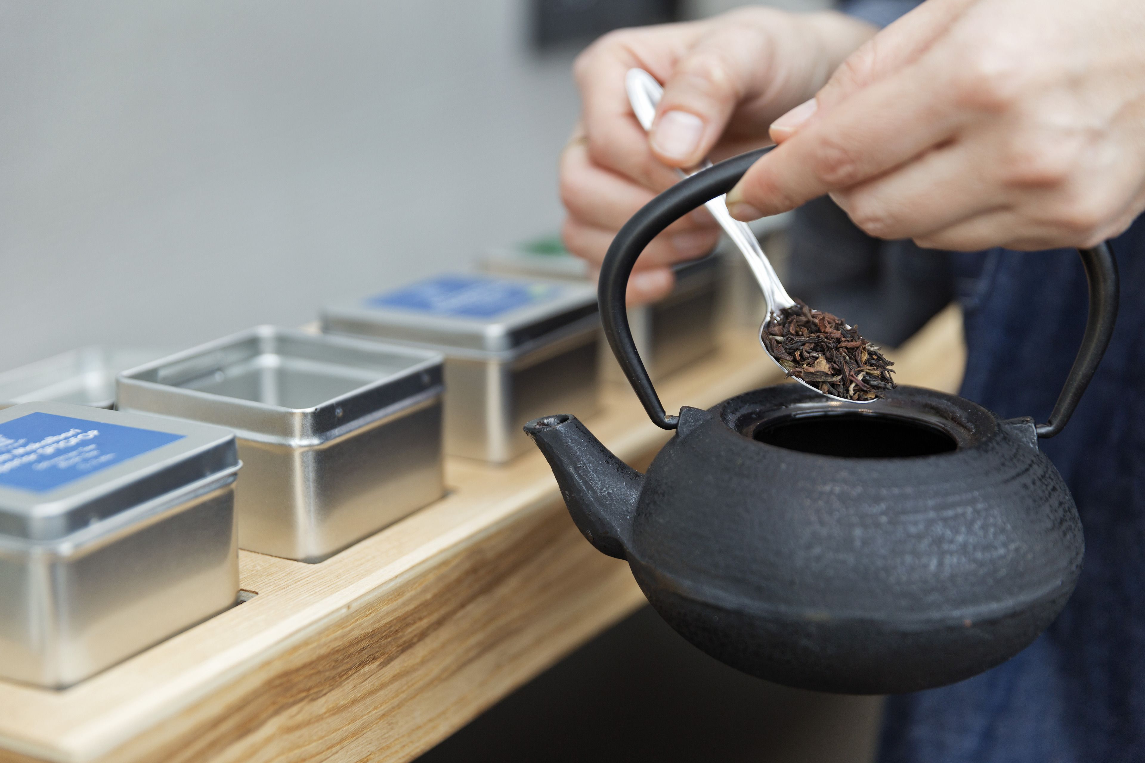 A person holding a tea kettle with a spoonful of tea leaves. There are metal containers with labels on a wooden surface beside the kettle.