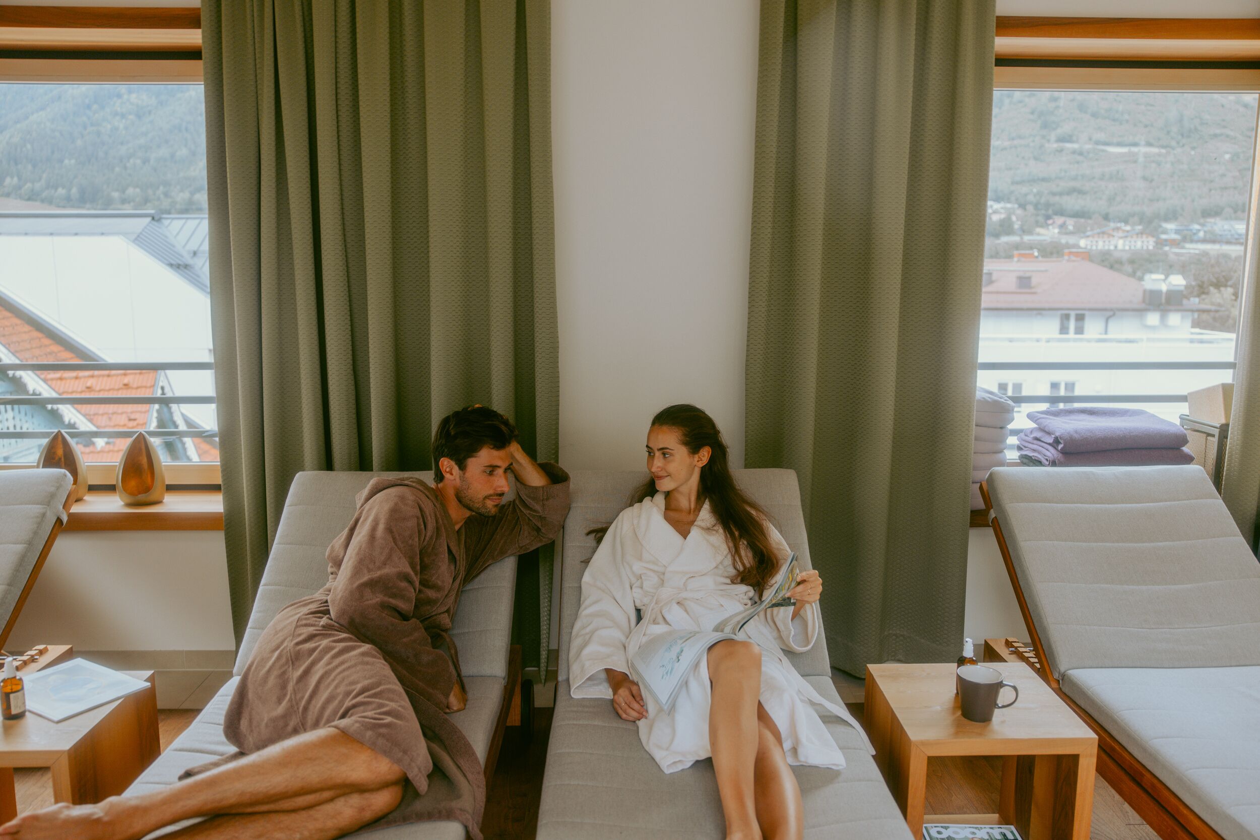 Ein Zimmer mit zwei Liegesesseln, einem Holztischchen mit einer Tasse und Blick auf eine gebirgige Landschaft durch große Fenster.