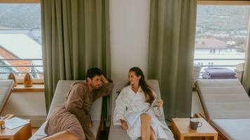 Ein Zimmer mit zwei Liegesesseln, einem Holztischchen mit einer Tasse und Blick auf eine gebirgige Landschaft durch große Fenster.