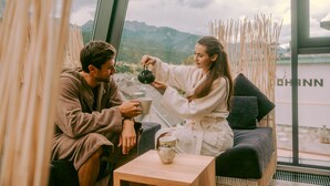 A couple sitting on a sofa with a view of mountains through a large window. - Stadthotel brunner (Schladming)