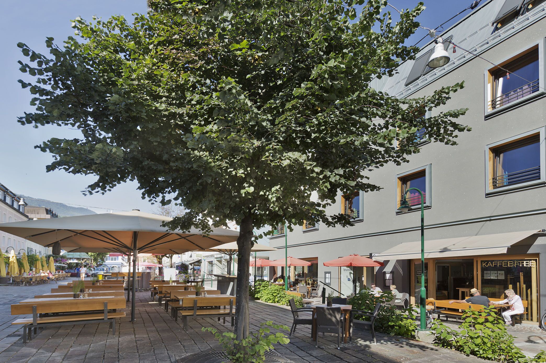 Ein Café mit Außenbereich, Holzbanken und Sonnenschirmen, ein Baum und ein Gebäude mit dem Schild 'KAFFEBAR'.