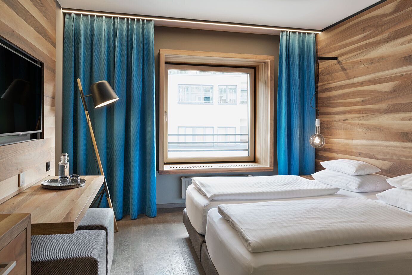 Habitación de hotel con pared de madera, un escritorio con lámpara, cama con almohadas y una ventana con cortinas.