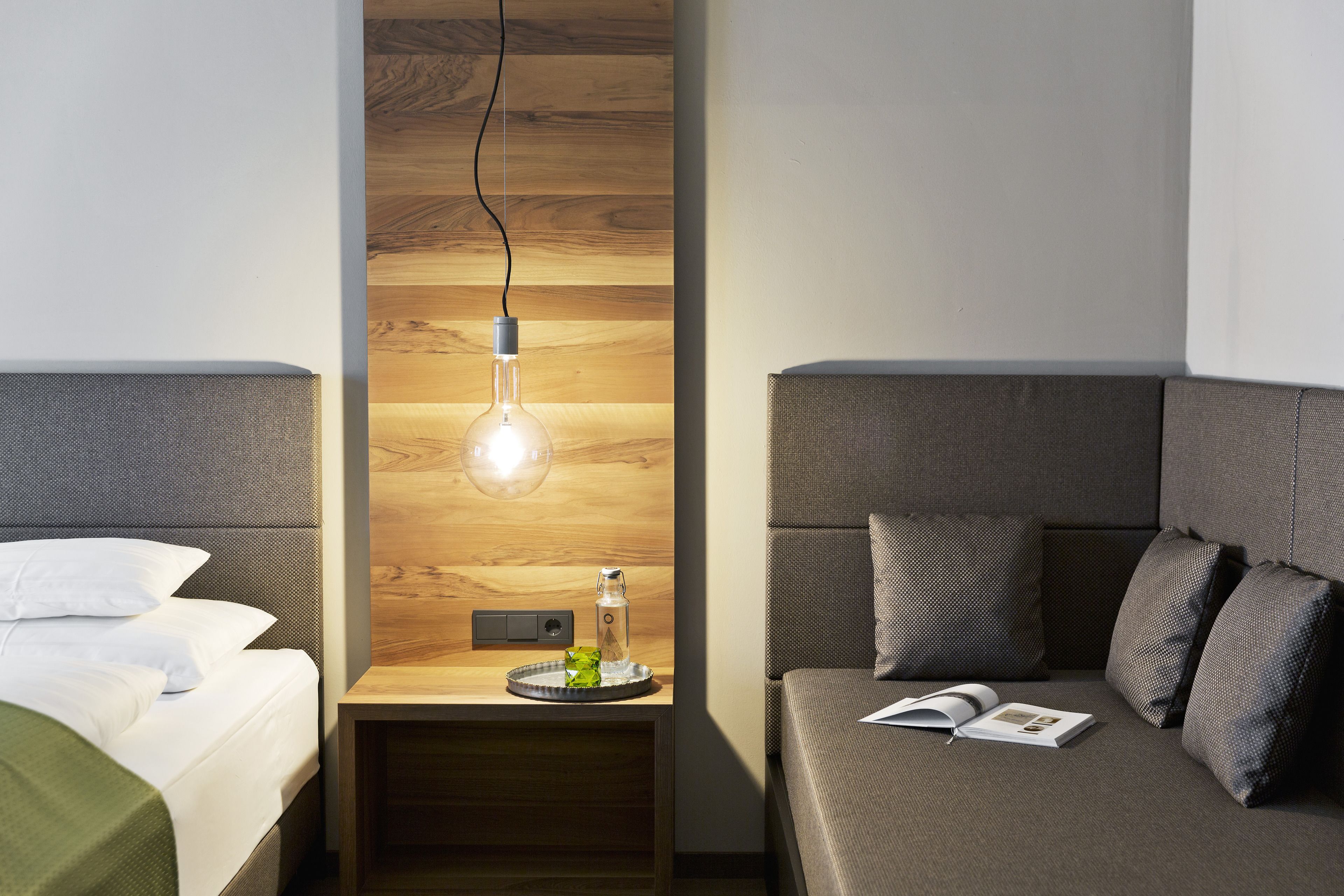 A modern hotel room with a wooden accent wall, a bedside table with a lamp, a bed with white linens, and a sofa with a book on it.