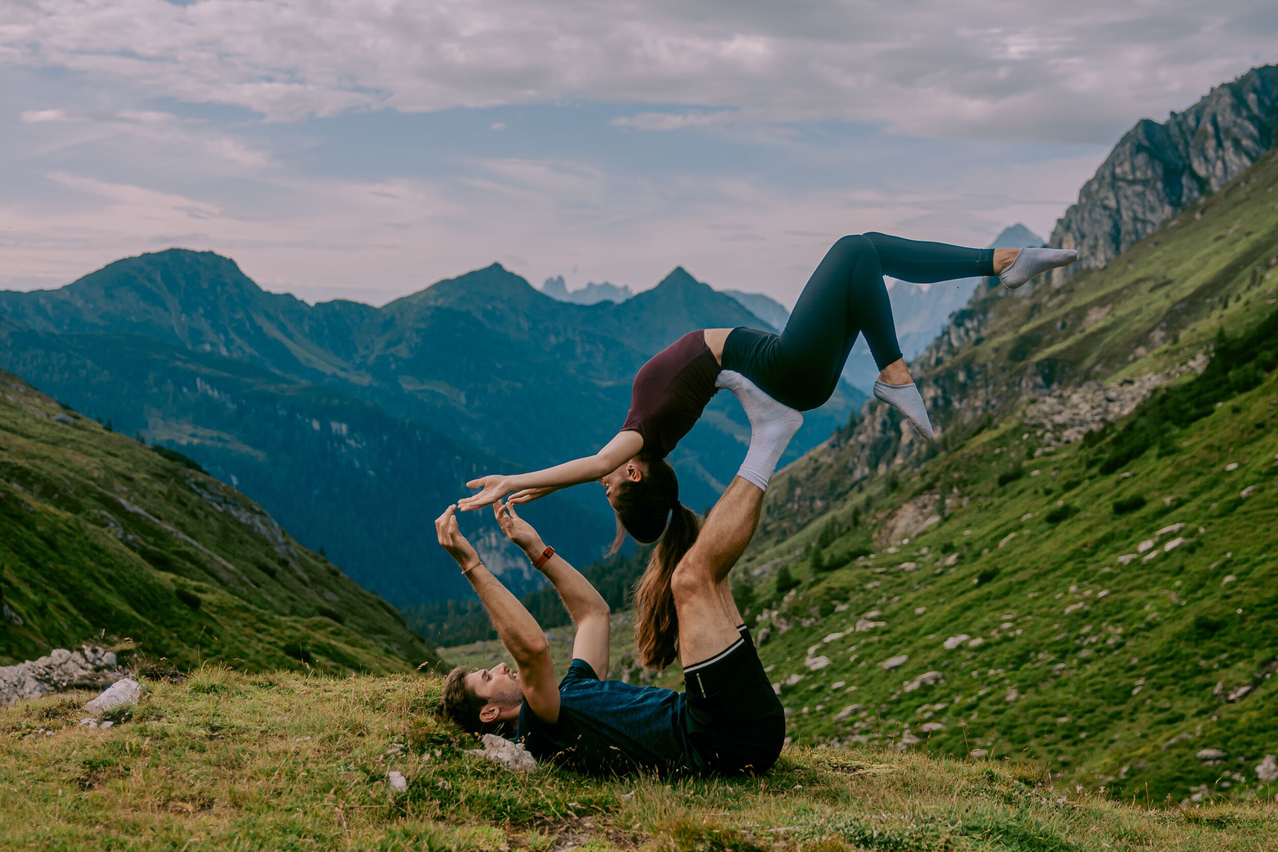 Two people performing an acrobatic move on a grassy hillside with mountains in the background.