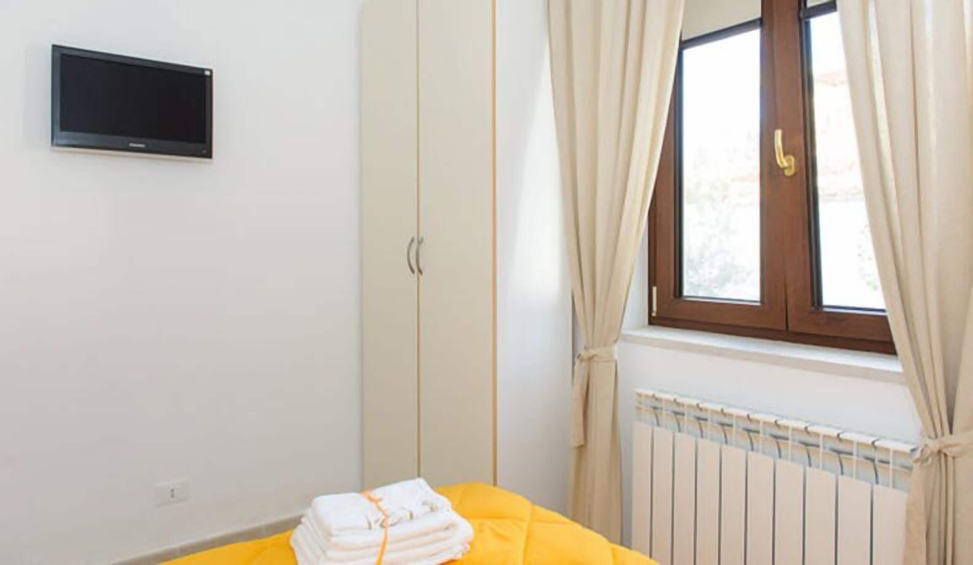 Een slaapkamer met een bed, een aan de muur bevestigde televisie, een raam met gordijnen en een radiator.
