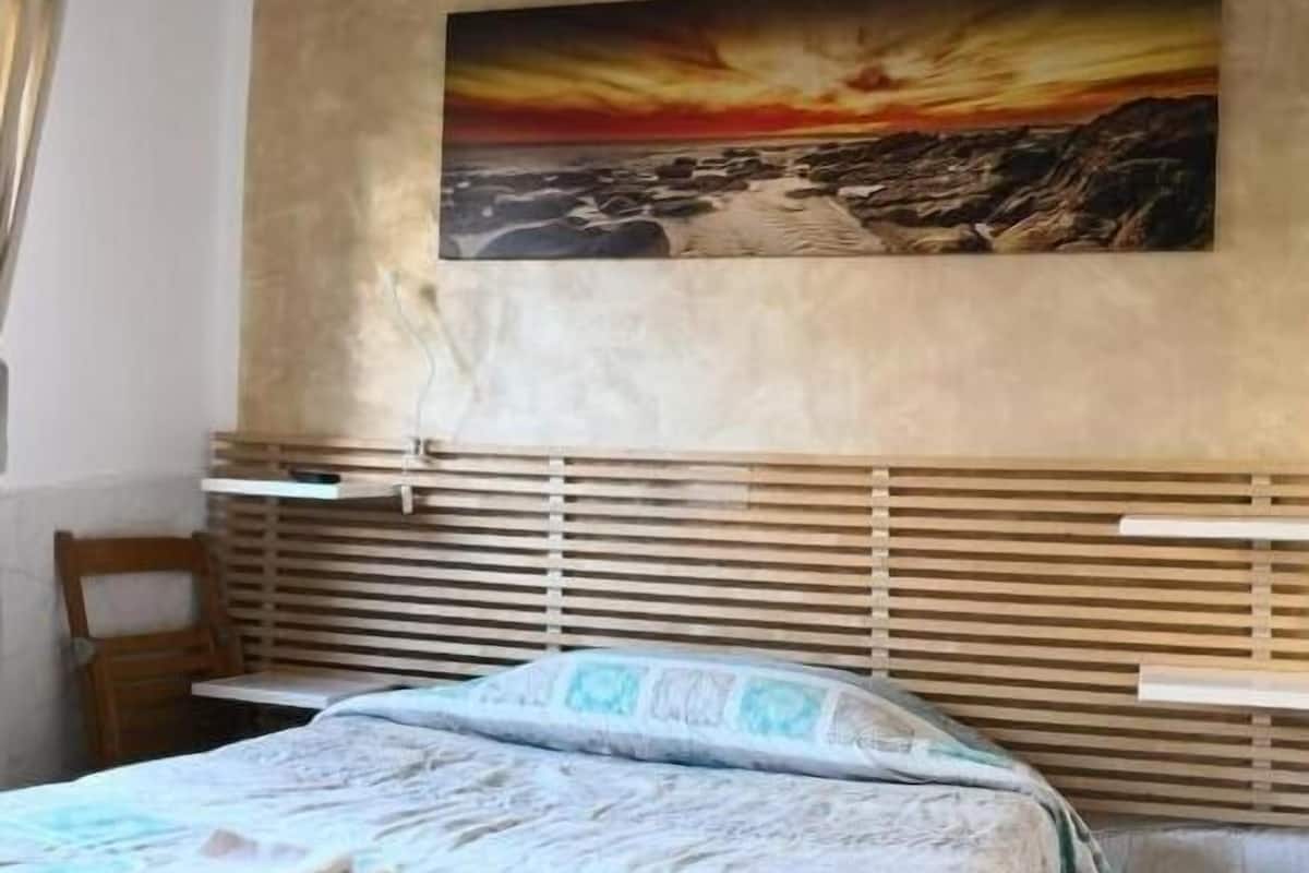 A bedroom with a bed, a wooden headboard, a painting of a sunset, and a window with blinds.