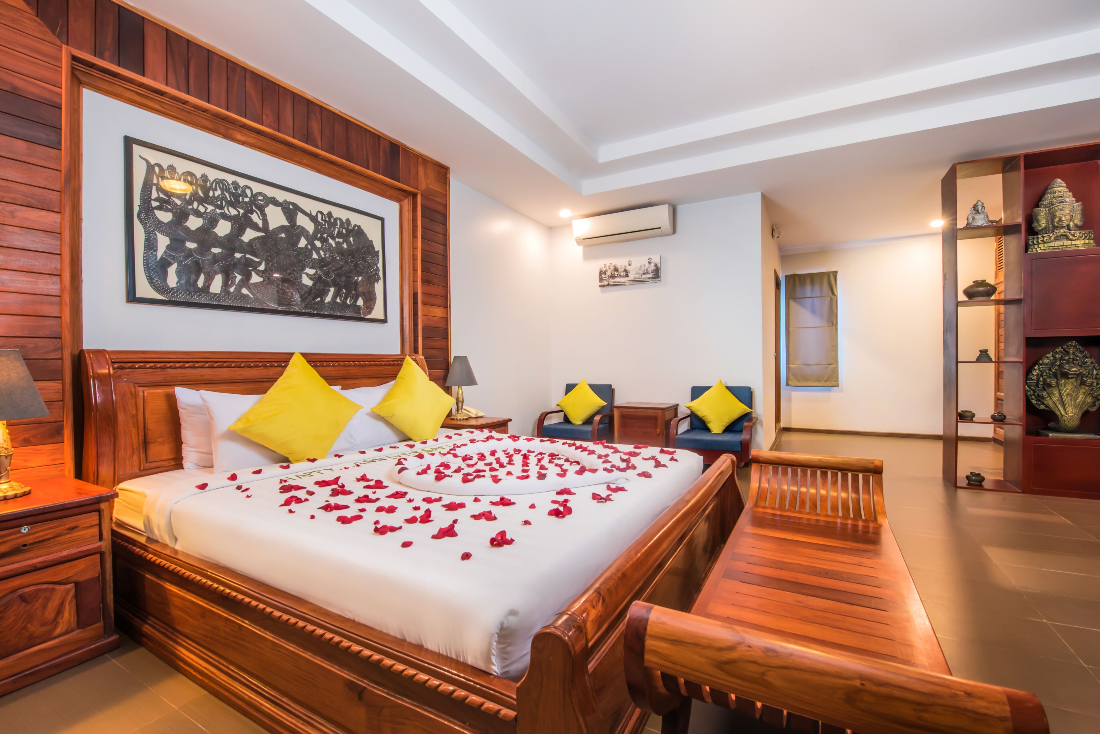 Una camera d'albergo con un letto di legno, una panca, un comodino, una lampada, un condizionatore d'aria a parete e uno scaffale con oggetti decorativi.