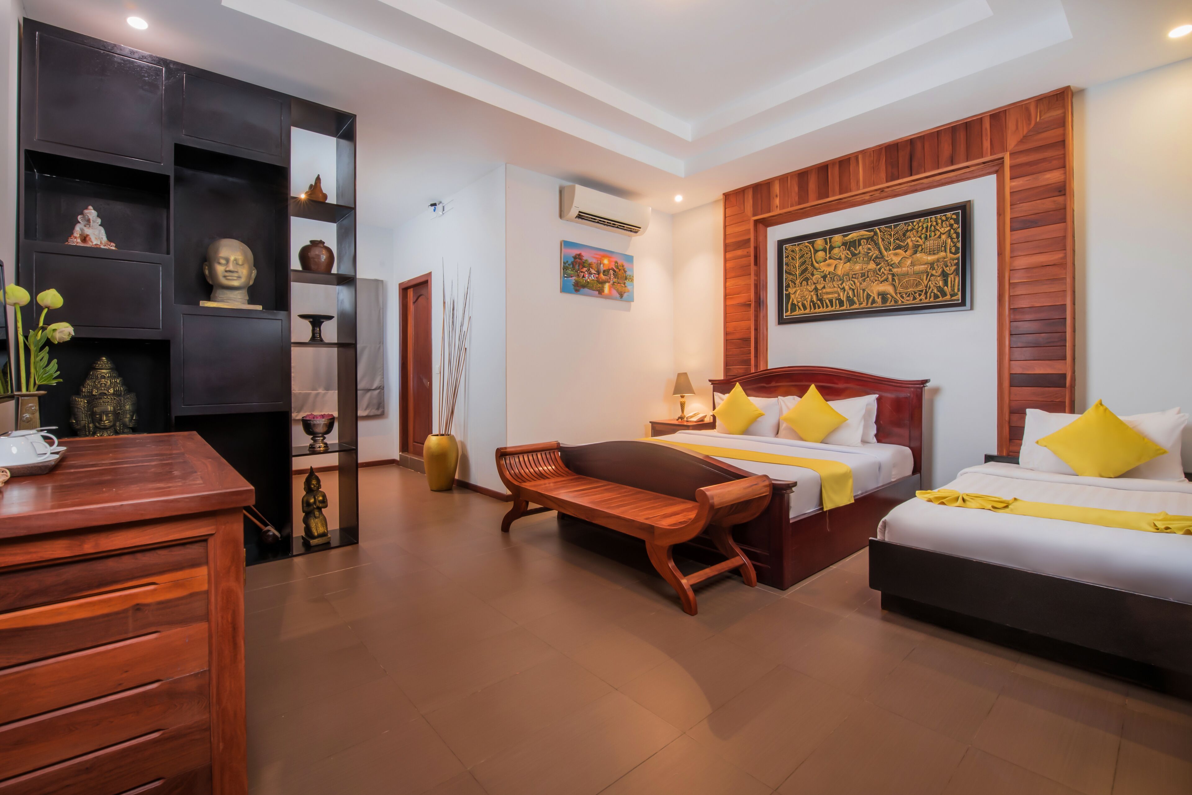 A hotel room with a wooden bench, two beds with yellow pillows, a wooden dresser, and shelves with decorative items.