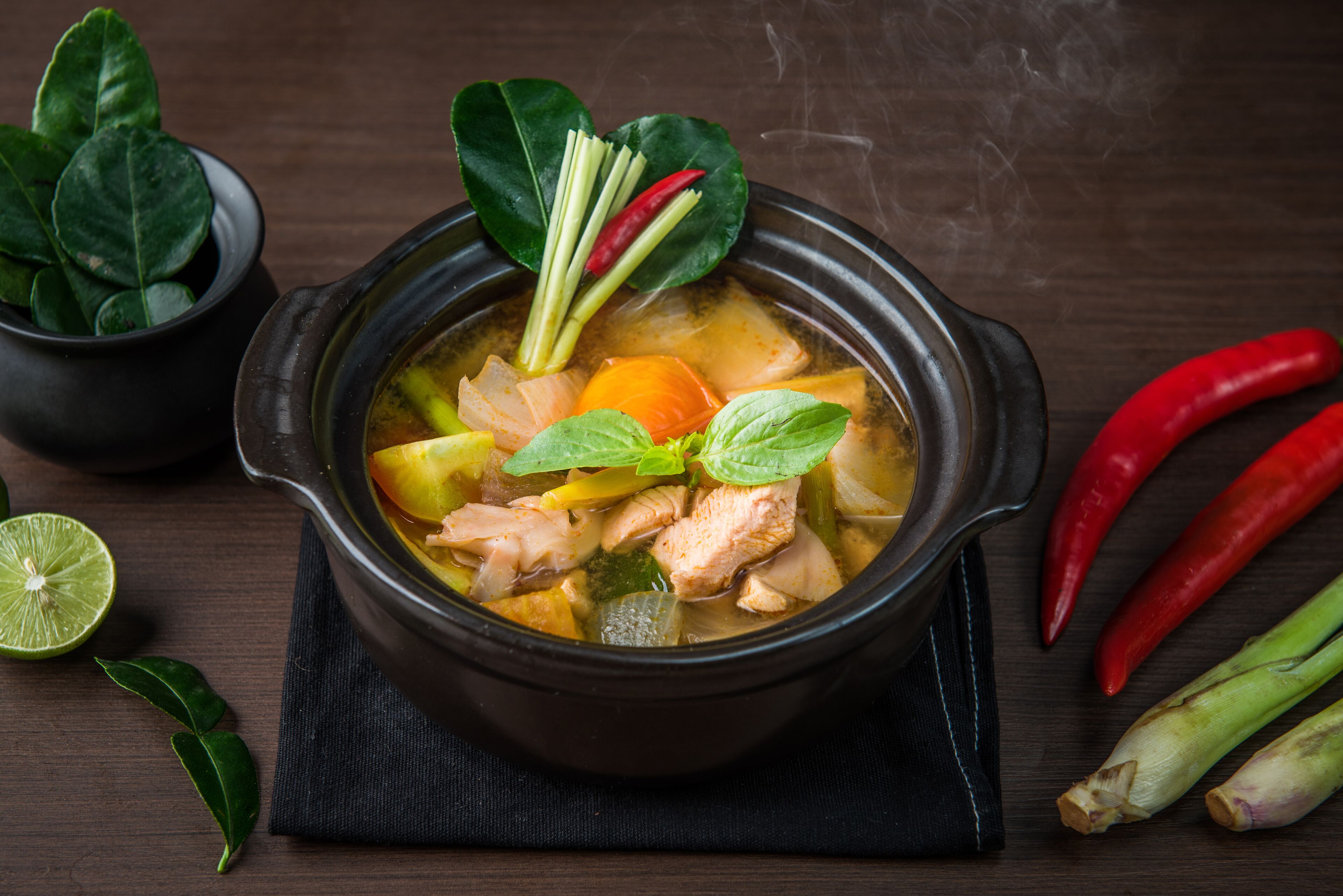 A bowl of soup with various vegetables and herbs, accompanied by a lime half and some chili peppers.