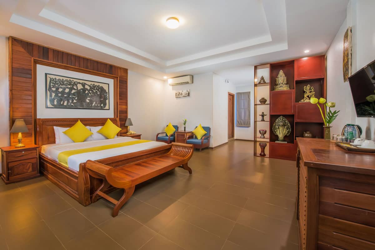 A spacious bedroom with a large bed, wooden headboard, and a bench. There is a desk with a chair, a shelf with decorative items, and a framed artwork on the wall.