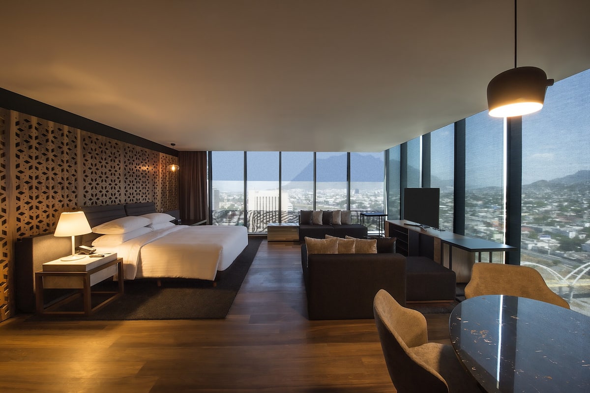 Une chambre d’hôtel moderne, dotée d’un grand lit, d’un canapé, d’une table et d’un téléviseur. La chambre dispose de grandes fenêtres offrant une vue sur la ville.