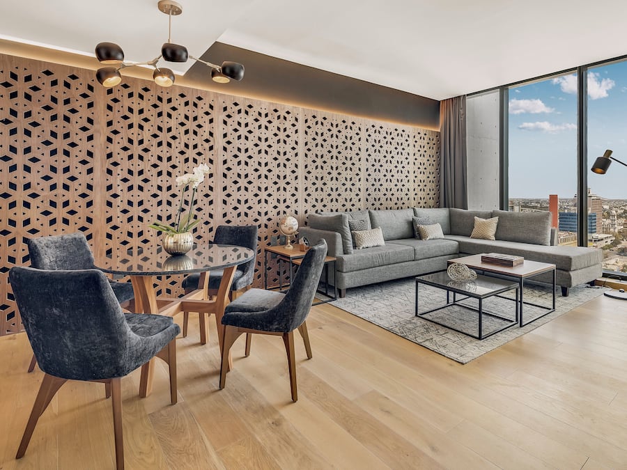 Una sala de estar moderna con un sofá gris, dos sillas oscuras, una mesa de centro redonda y piso de madera. Se aprecia un panel decorativo y un amplio ventanal con vista a la ciudad.