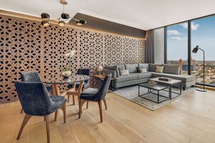 Una sala de estar moderna con un sofá gris, dos sillas oscuras, una mesa de centro redonda y piso de madera. Se aprecia un panel decorativo y un amplio ventanal con vista a la ciudad.