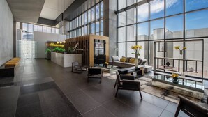 Een ruime lobby met een modern ontwerp, met een zithoek met banken en stoelen, een receptiebalie en grote ramen met uitzicht op de lucht en een gebouw.