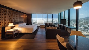 Een moderne hotelkamer met een groot bed, een bank, een eettafel en een televisie. De kamer heeft grote ramen met uitzicht over de stad.