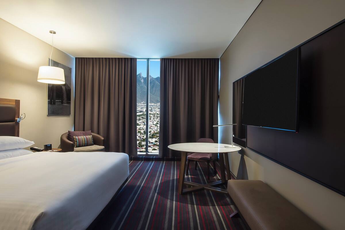 Une chambre d’hôtel avec un grand lit, un bureau avec une chaise, un téléviseur fixé au mur et une vue sur les montagnes par la fenêtre.