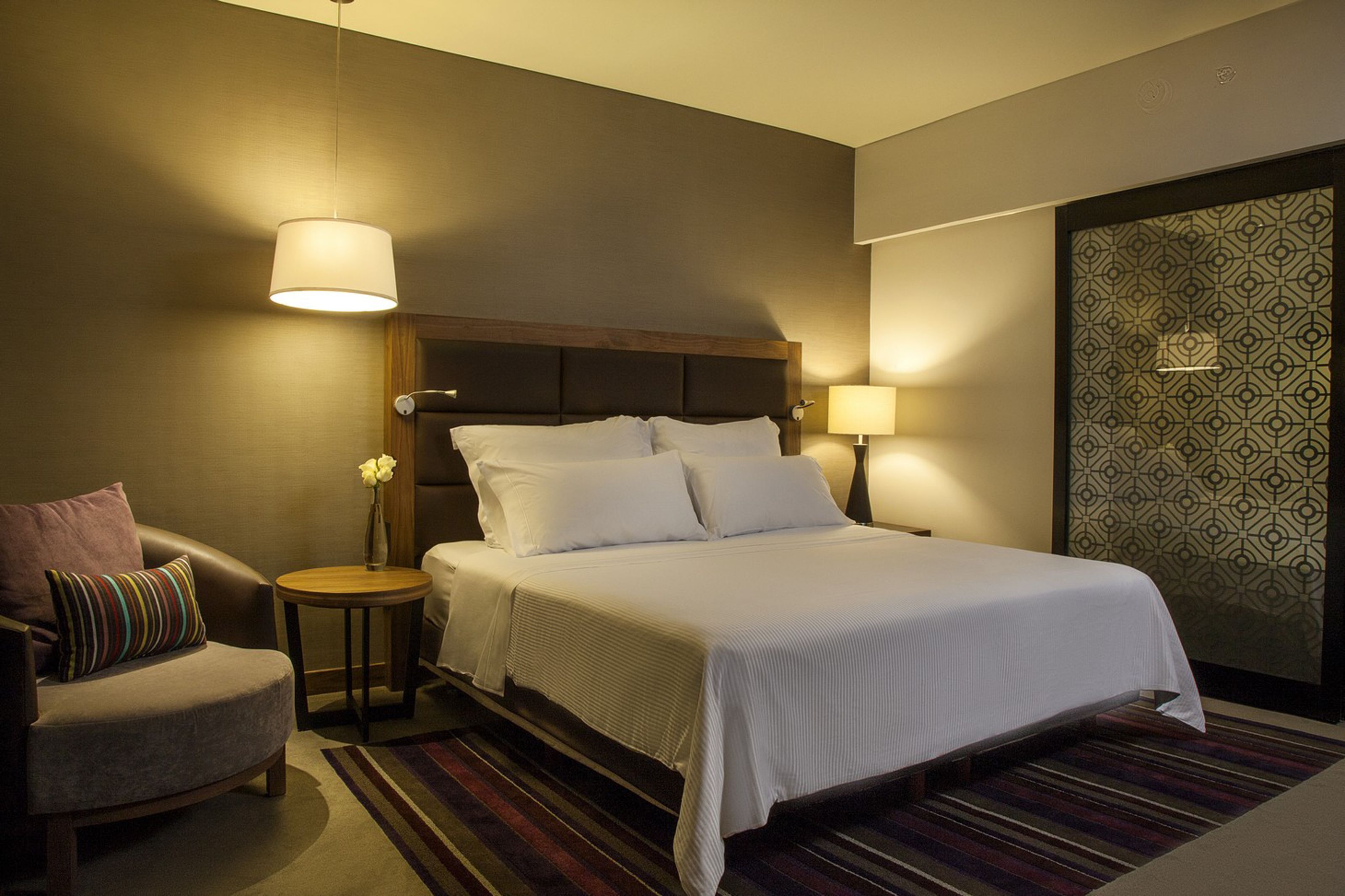 A hotel room with a bed, a chair, a nightstand, and a wall with a geometric pattern.