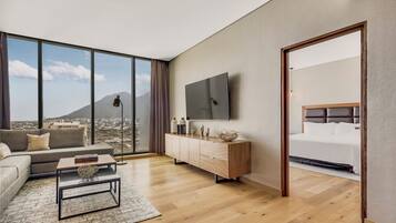 A modern hotel room with a large window, a flat-screen TV, a sofa, a coffee table, and a wooden floor.