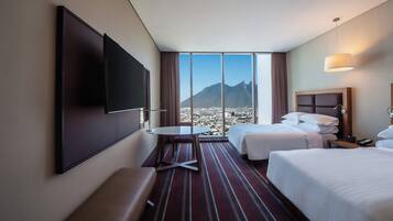 Quarto de hotel com duas camas, televisão, uma mesinha e vista para uma montanha.