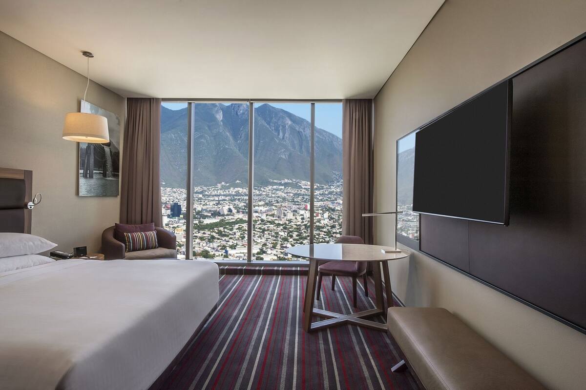 Une chambre d’hôtel avec un grand lit, un bureau, une chaise, une télévision et une vue sur la ville et les montagnes.