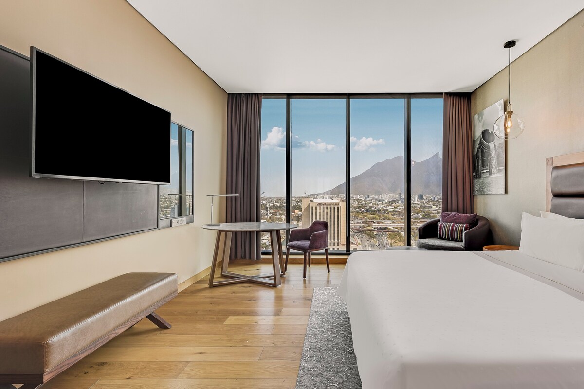 Een moderne hotelkamer met een groot bed, een flatscreen televisie, een klein tafeltje, een bankje en uitzicht op een stadsbeeld en bergen.