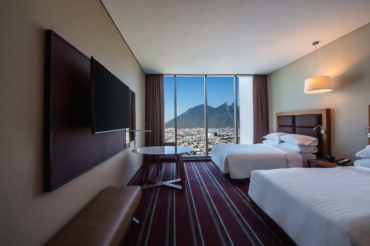 Une chambre d’hôtel avec une grande télévision, deux lits, une petite table et une vue sur la montagne.