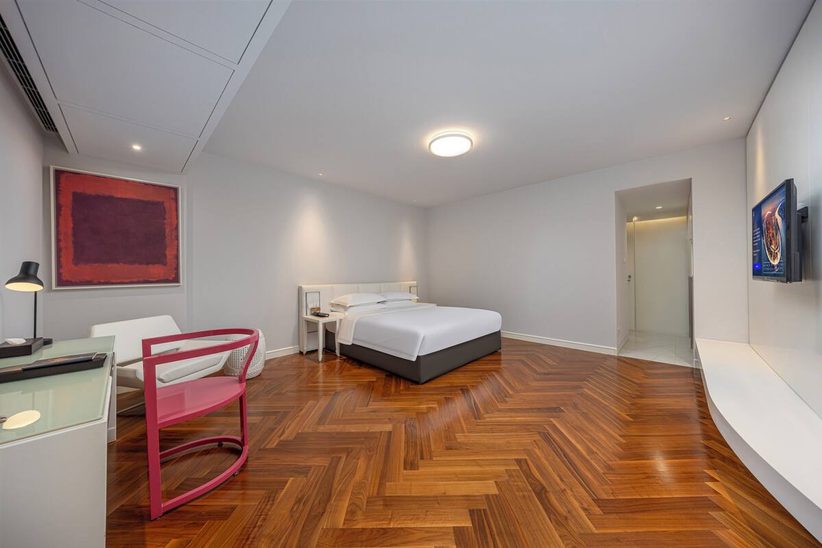 Ein modernes Hotelzimmer mit Holzdielen im Fischgrätmuster, einem Bett mit weißem Kopfteil, einem Schreibtisch mit Computer, einem Stuhl und einem großen abstrakten Gemälde an der Wand.