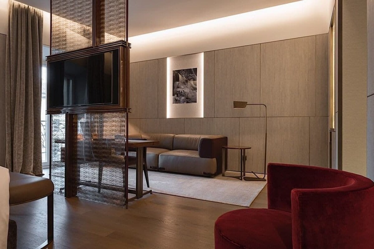 A modern hotel room with a bed, a red armchair, a sofa, a TV, and artwork on the walls.