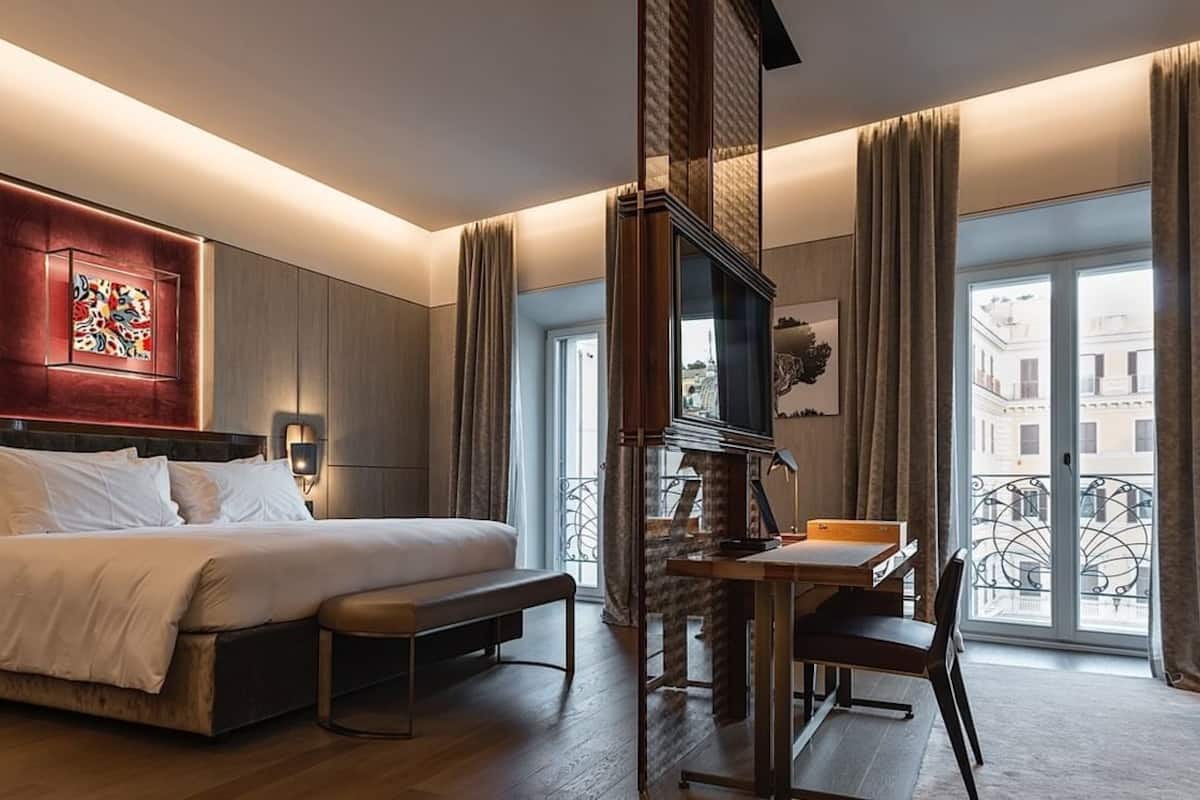A hotel room with a large bed, a desk, a chair, a TV, and a balcony with a view of buildings.