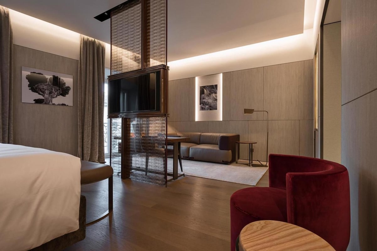 A modern hotel room with a bed, a TV, a sofa, a red chair, and a small table.