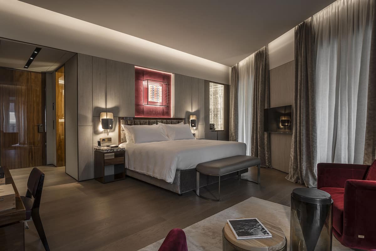 A modern hotel room with a large bed, bedside tables, a desk, a chair, and a TV.