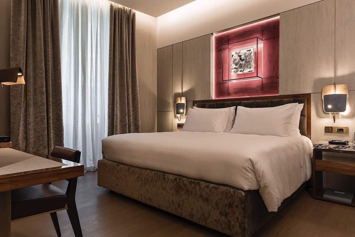 A hotel room with a large bed, a desk, a chair, a nightstand with a lamp, and a wall-mounted artwork.
