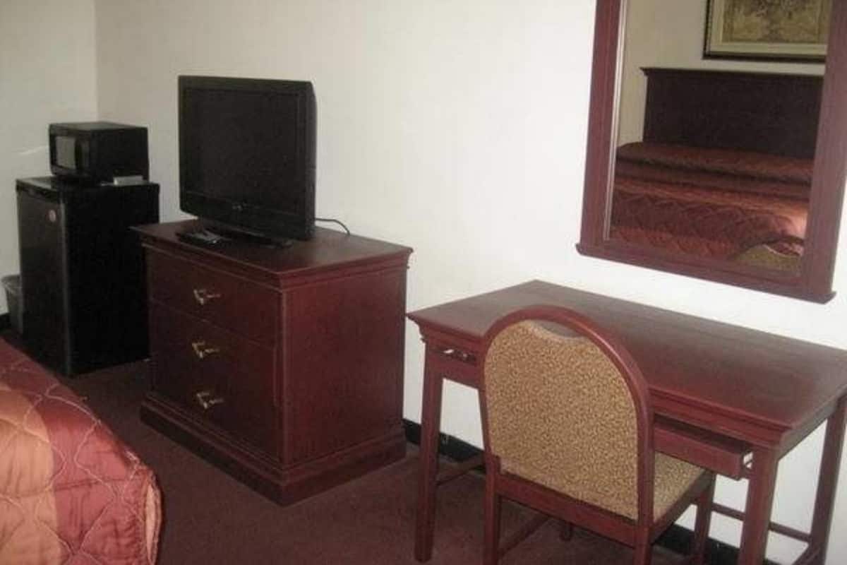Quarto de hotel com uma secretária de madeira, televisão sobre um móvel, uma cadeira e uma cama com cobertor vermelho.