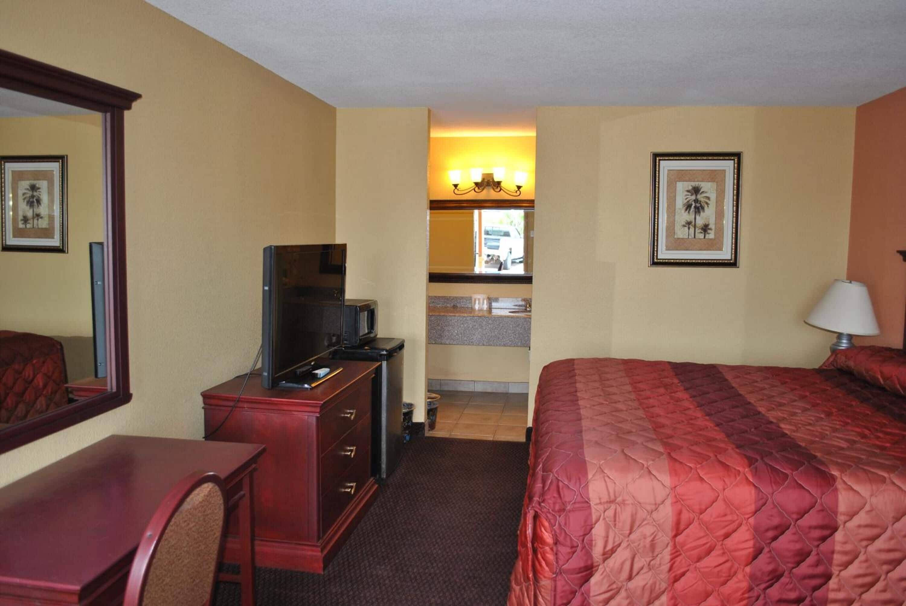Habitación de hotel con cama, escritorio, cómoda, televisor, espejo y kitchenette.