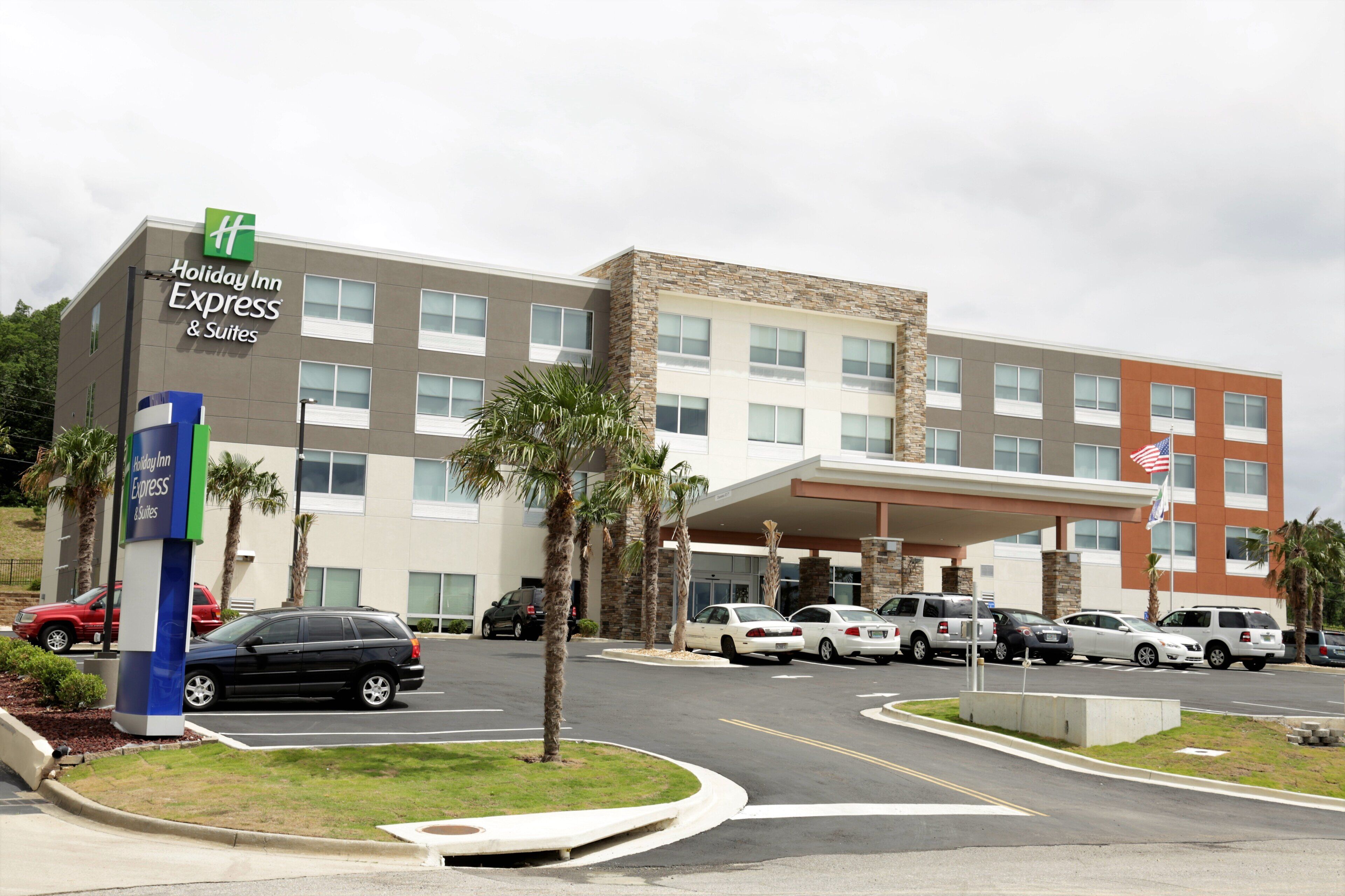 Un bâtiment de la chaîne Holiday Inn Express & Suites, doté d’un design moderne, avec une façade combinant pierre et verre, un parking où sont stationnées plusieurs voitures, et un mât sur lequel flotte le drapeau américain.