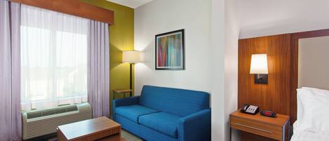 A hotel room with a blue sofa, a small wooden table, a bedside table with a lamp, a bed, and a window with curtains.