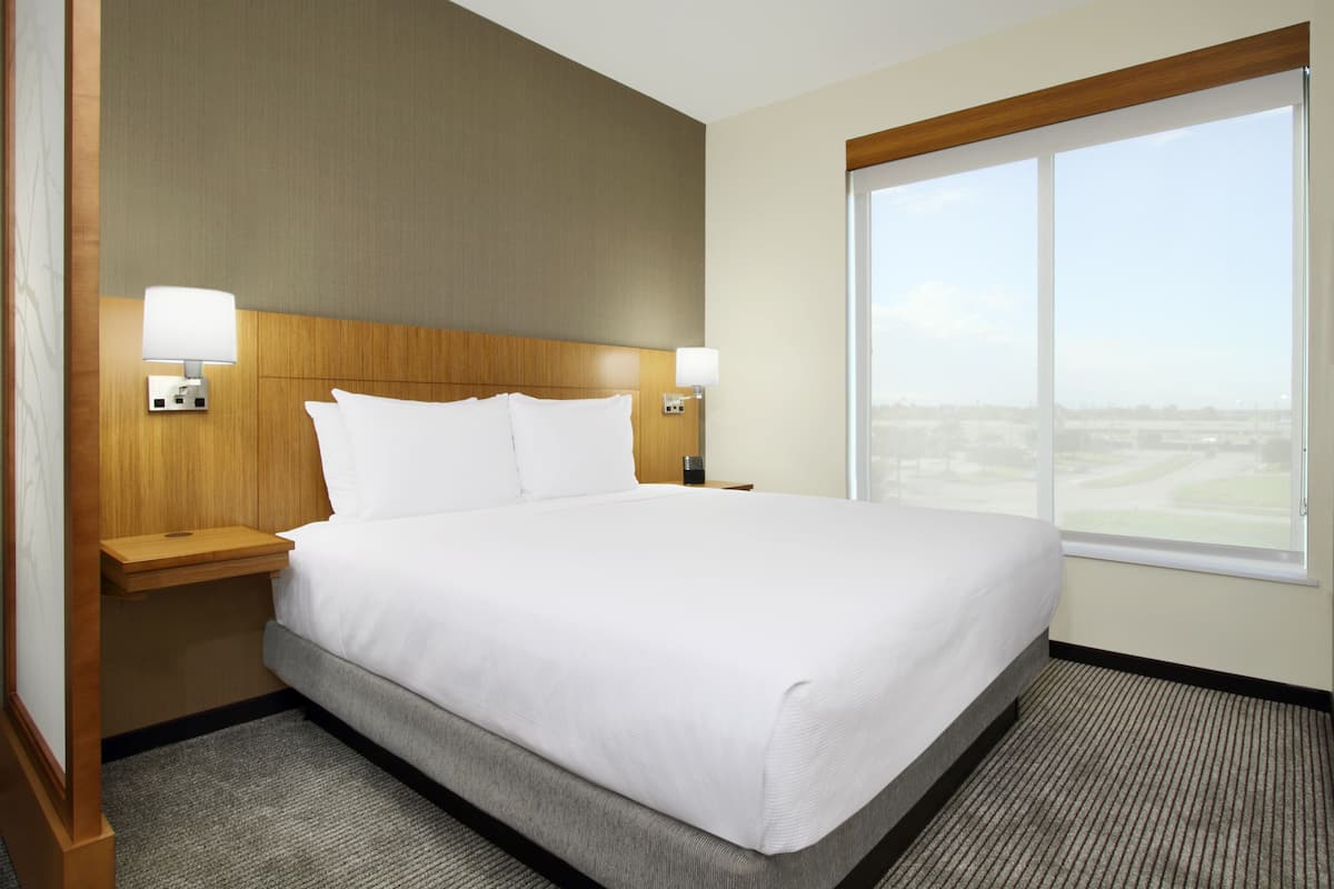A hotel room with a large bed, two bedside tables with lamps, a wooden headboard, and a large window offering a view of the city.