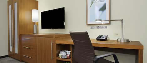 Habitación de hotel con un escritorio de madera, televisor, lámpara, silla, teléfono y estante con artículos de aseo.