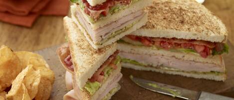 A sandwich cut in half with visible layers of meat, lettuce, and tomato, accompanied by potato chips and a knife on a wooden board.