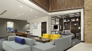 Un hall d’hôtel moderne avec un canapé d’angle gris, des fauteuils jaunes, une table basse en bois et un téléviseur fixé au mur.