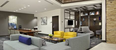 A modern hotel lobby with a grey sectional sofa, yellow armchairs, a wooden coffee table, and a TV on a wall.