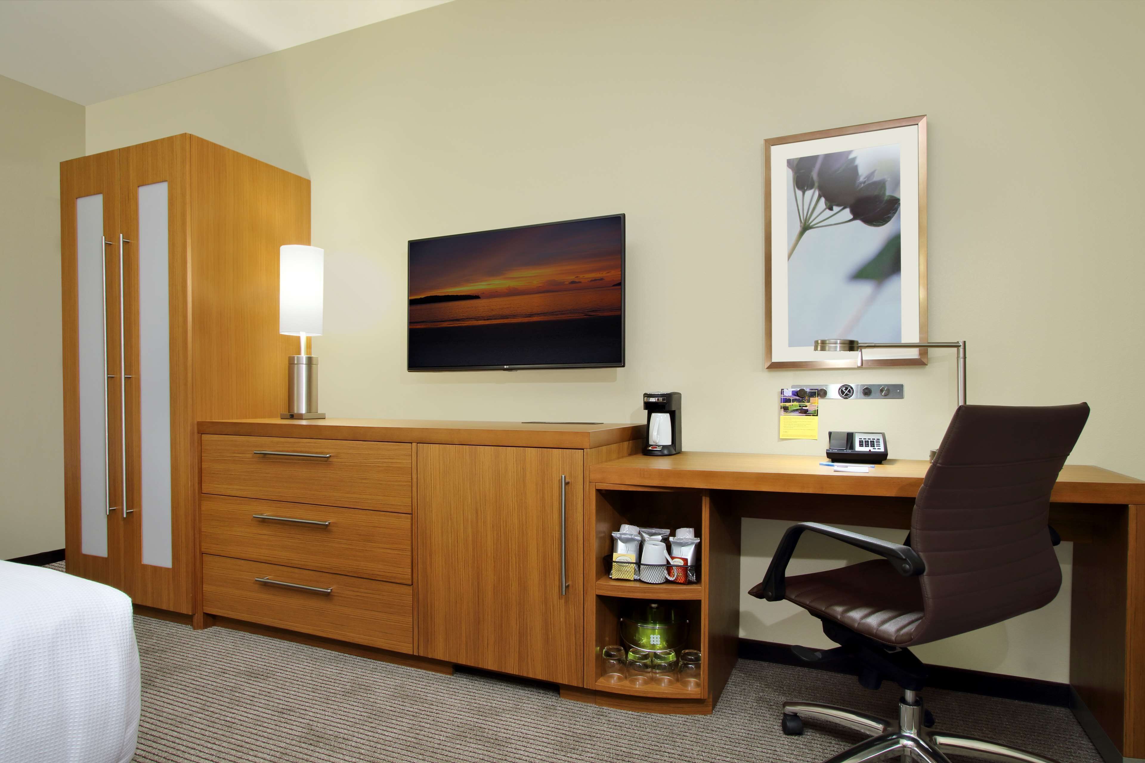 Habitación de hotel con escritorio de madera, televisor, un cuadro enmarco, cafetera y una silla.