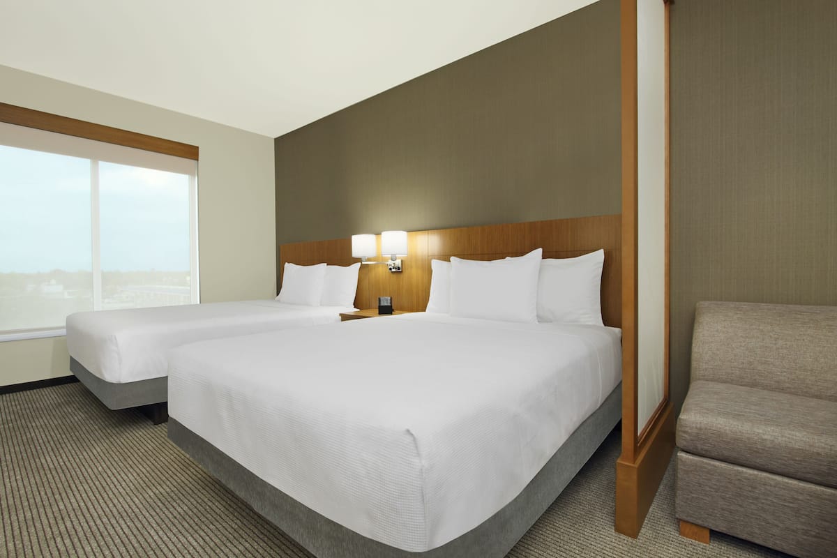 A hotel room with two beds, a nightstand with lamps, a view of the city, and a large window.