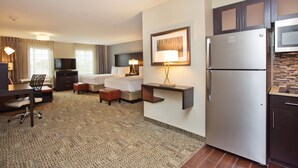 Quarto de hotel com cama, escrivaninha, cadeira e cozinha compacta, incluindo frigorífico e micro-ondas.