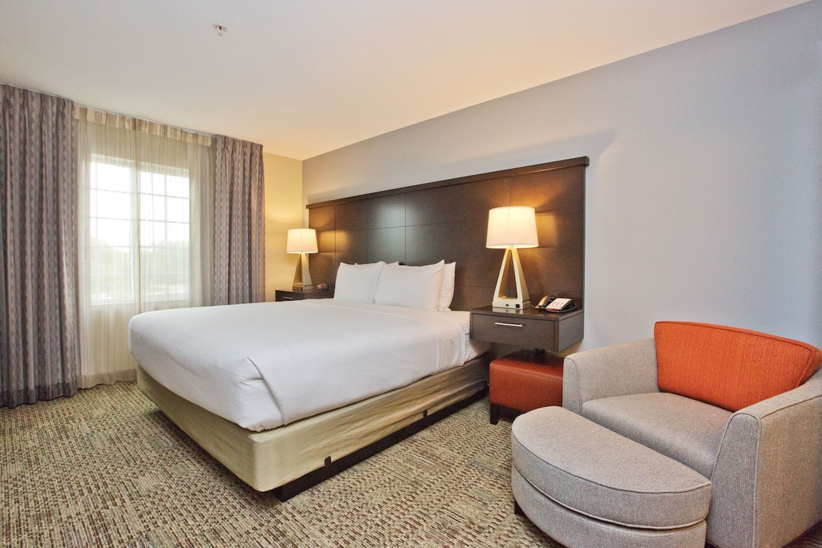 Una habitación de hotel con una cama grande, dos lámparas de noche, una mesita de noche, un sillón rojo y gris y una ventana con cortinas.