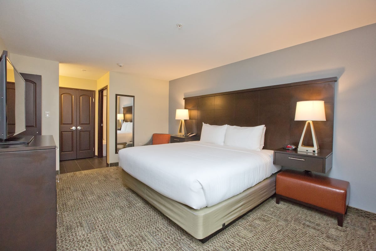 Una habitación de hotel con una cama grande, dos mesitas de noche con lámparas, un banco de madera, un aparador y una puerta que da a otra habitación.