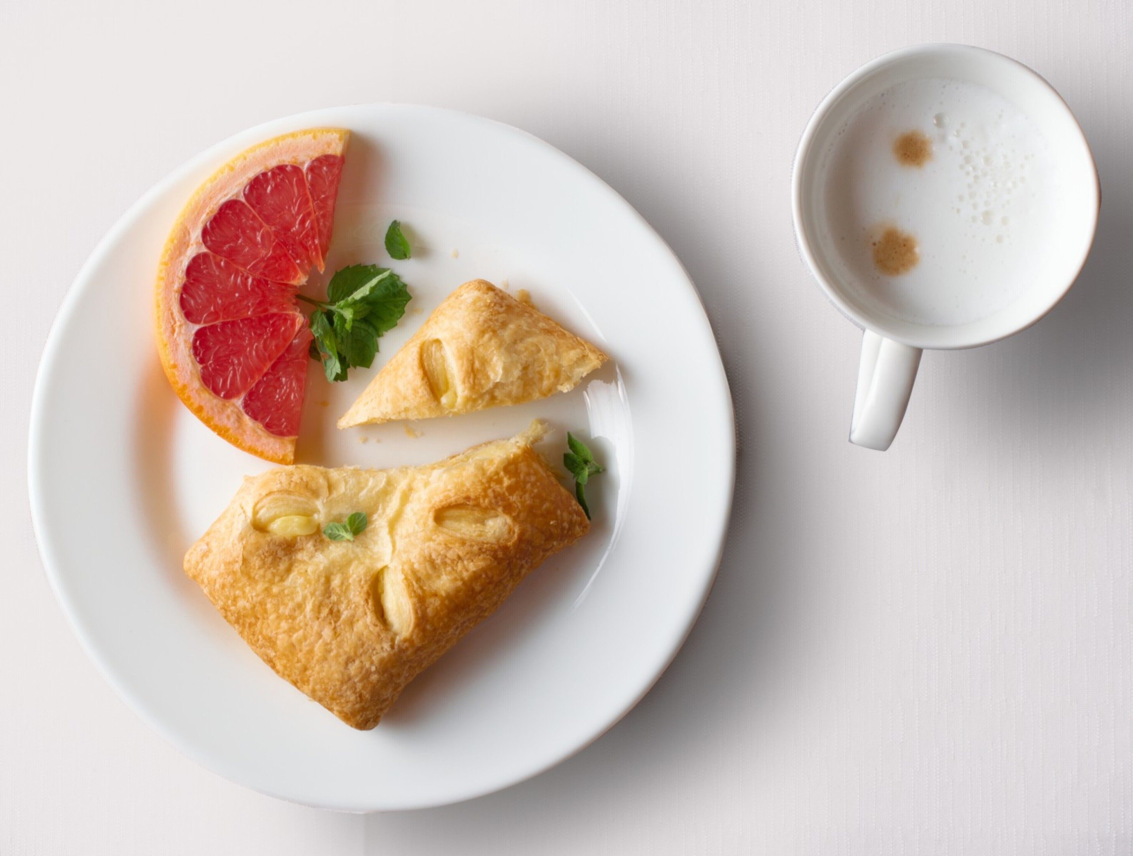 Un piatto con due pasticcini e una fetta di pompelmo, e una tazza di caffè su una superficie bianca.